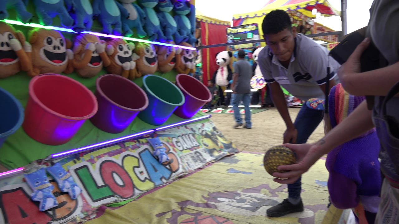 A young carnival attendant stands by the ball toss booth while an adult hand holds a ball near the angled row of buckets in San Jose Del Cabo.