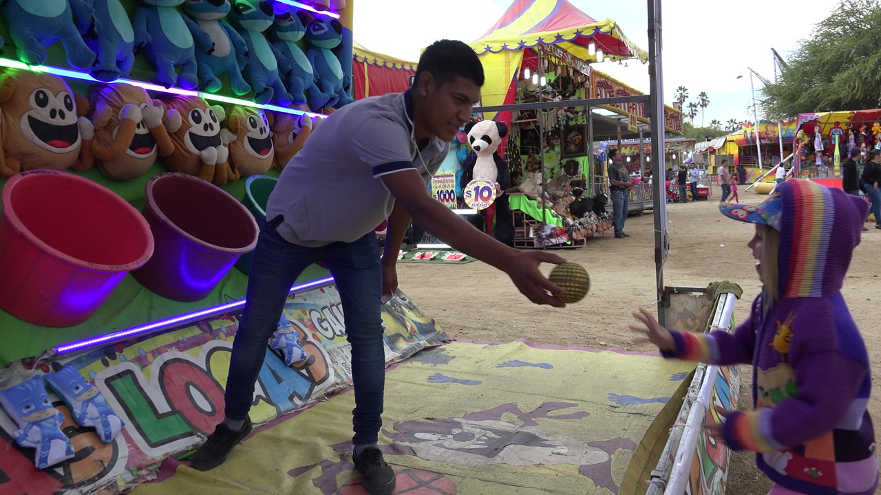 In San Jose del Cabo, Mexico, the child stands closer to the buckets while the adult holds a ball over the booth mat, with plush monkeys and bright tent tops behind them.