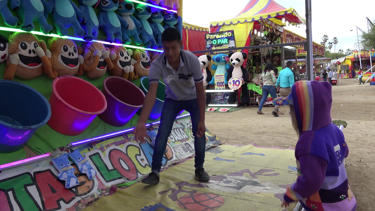 In San Jose del Cabo, Mexico, a child in a purple hoodie faces a carnival game booth while a young attendant bends near colored tubs and stuffed prizes.