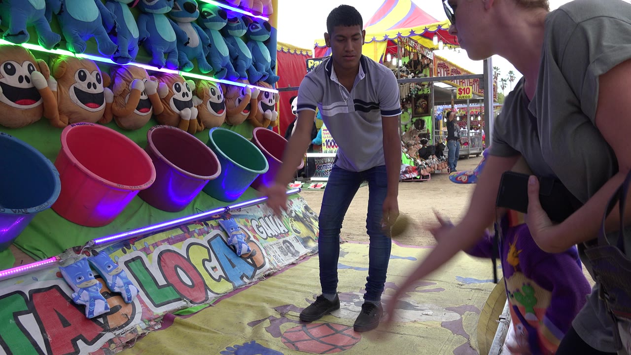 At a San Jose del Cabo carnival game booth, a male attendant stands by colorful toss buckets while an adult player leans in from the right edge.
