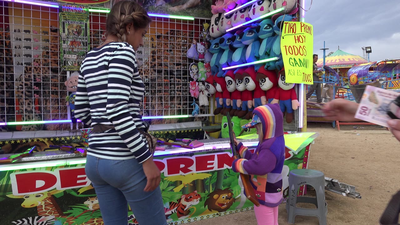 A woman in a striped shirt stands beside a small child in a purple hooded sweater at a prize shooting booth in San Jose Del Cabo, Mexico.