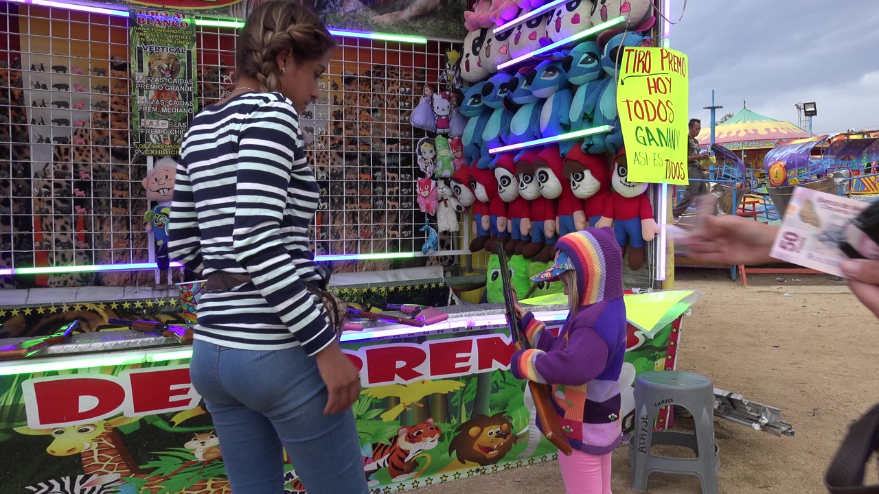 An adult hand holding cash enters the frame while the woman and child remain beside the carnival shooting game in San Jose Del Cabo, Mexico.
