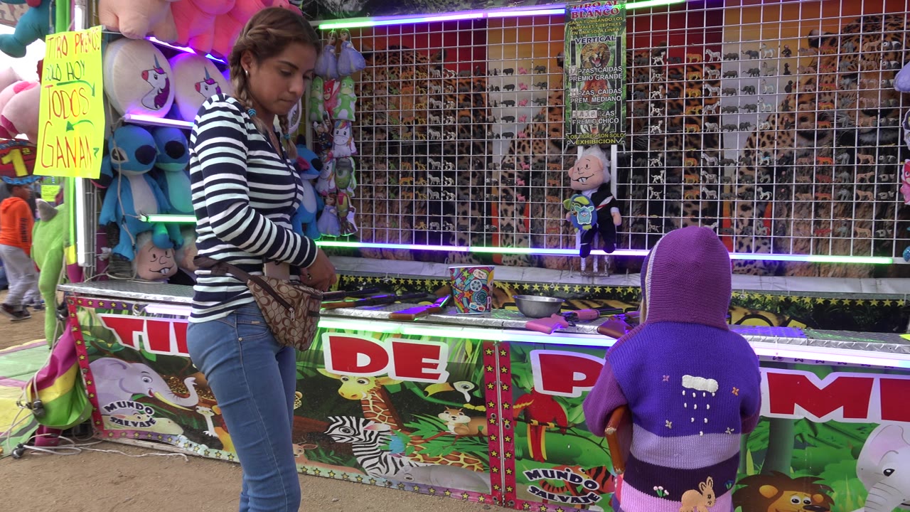 A woman stands beside a small child at a colorful fair prize booth in San Jose del Cabo, Mexico, with plush toys, neon strips, and target panels behind the counter.