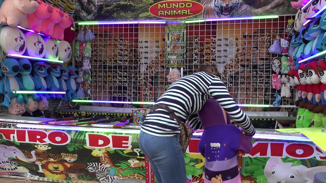 In San Jose del Cabo, Mexico, a woman in a striped shirt leans over a child in a purple seat at a shooting gallery booth with plush prizes and neon strips behind them.