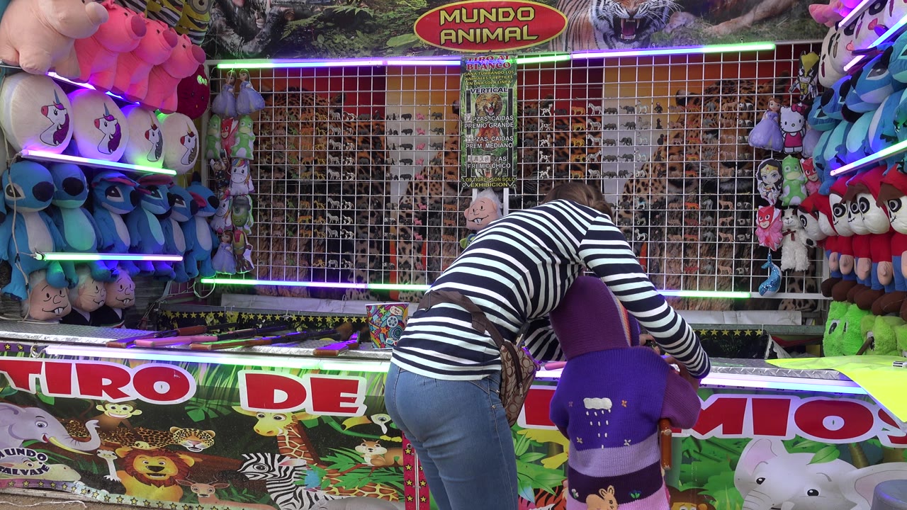 The woman remains bent toward the child at the San Jose del Cabo fair booth, with a purple chair, toy rifle, stuffed toys, and a gridded target wall visible around them.