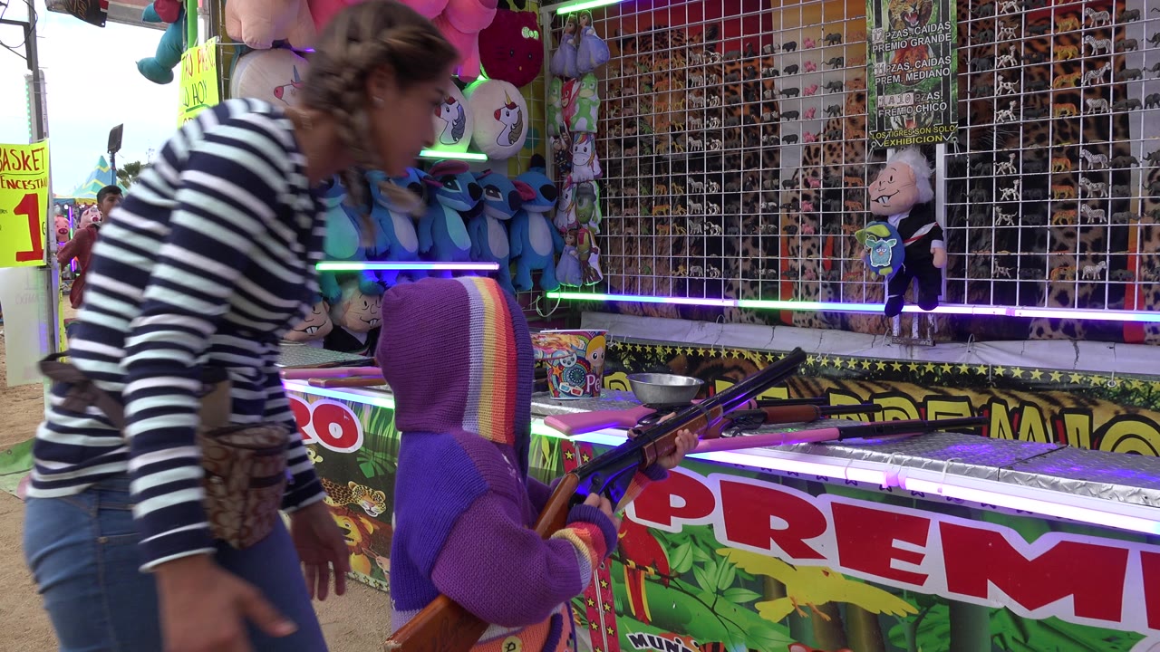 Woman bends beside a child at a fair game counter with toy rifles and prizes.