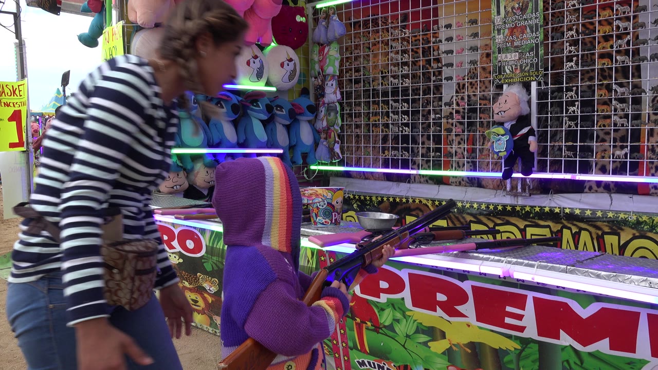 In San Jose Del Cabo, Mexico, the woman remains close to the child as the child holds a toy rifle near a counter lined with bright signs and stuffed prizes.