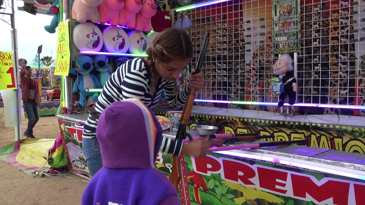 In San Jose Del Cabo, Mexico, a young woman in a striped shirt leans over a child in a purple hoodie beside a toy rifle at a colorful prize booth.