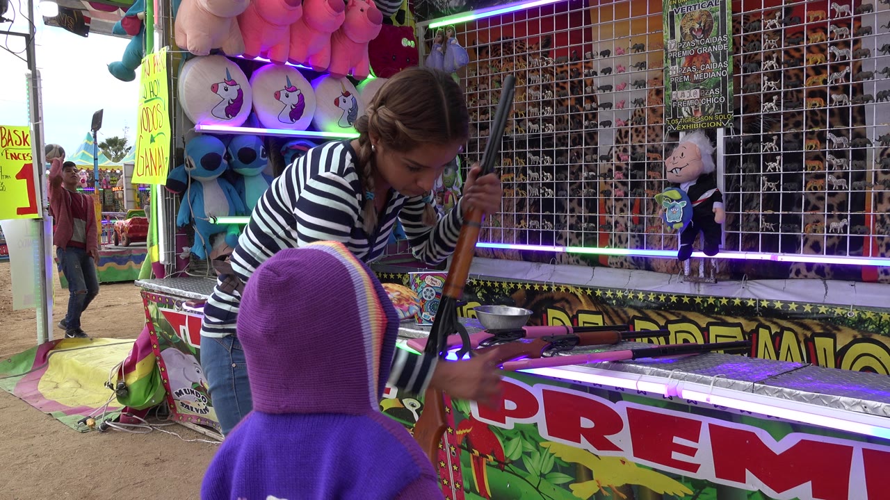 The woman bends forward and reaches toward the toy rifle while the child faces the carnival counter, with plush toys and gridded prize shelves behind them in San Jose Del Cabo, Mexico.