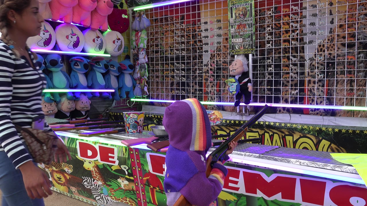 At a San Jose del Cabo carnival booth, a woman in a striped shirt passes beside a child in a purple hood holding a toy rifle near plush prizes and a target wall.
