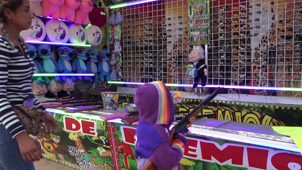 The adult worker moves closer to the child at the San Jose del Cabo fair game, with stuffed toys on lit shelves and a gridded target board behind them.