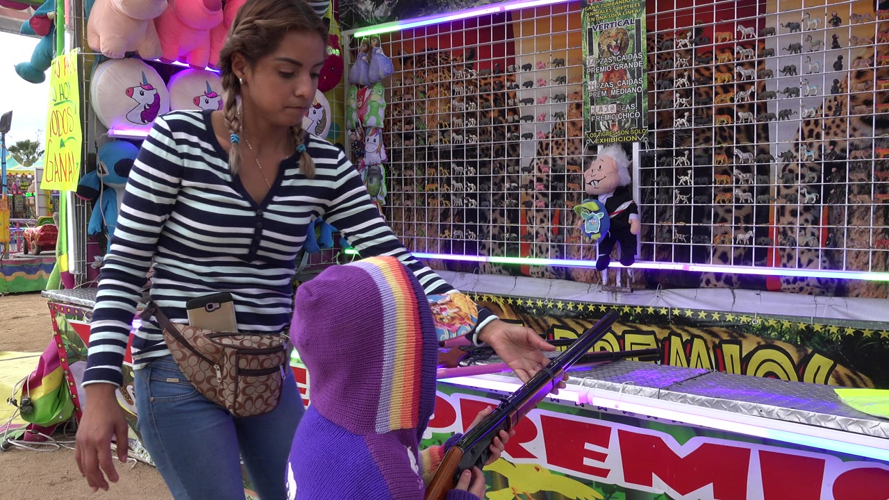 At 0:01 in San Jose Del Cabo, Mexico, the child keeps the toy rifle pointed toward the booth target area as the adult woman remains beside the counter under colored lights.