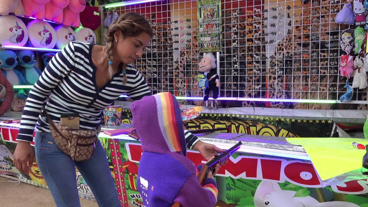 At a San Jose del Cabo outdoor game booth, a child in a purple hood faces a toy rifle while a woman in a striped shirt stands beside colorful prizes and target racks.