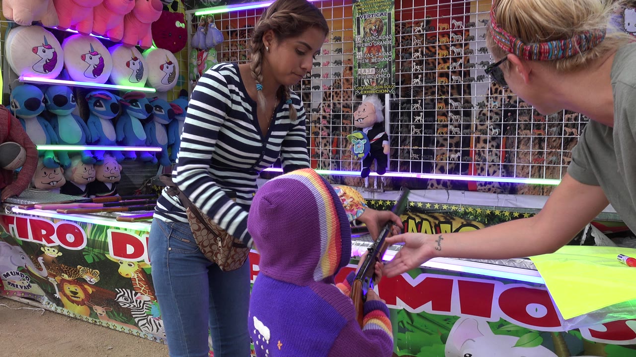 In San Jose del Cabo, Mexico, a child in a purple hood faces a carnival shooting game while an adult reaches in and a striped-shirt attendant stands beside plush prizes.