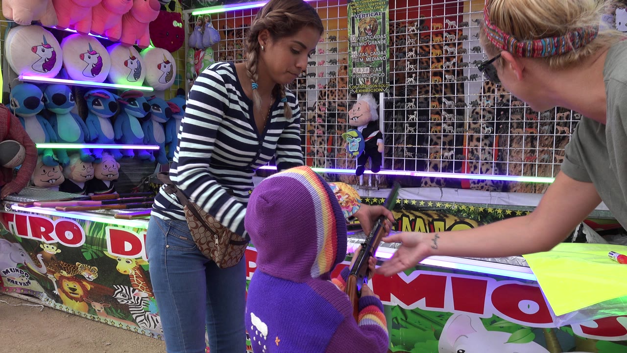 An adult bends toward a child holding a toy rifle at the San Jose del Cabo carnival booth while the attendant watches from behind the counter.