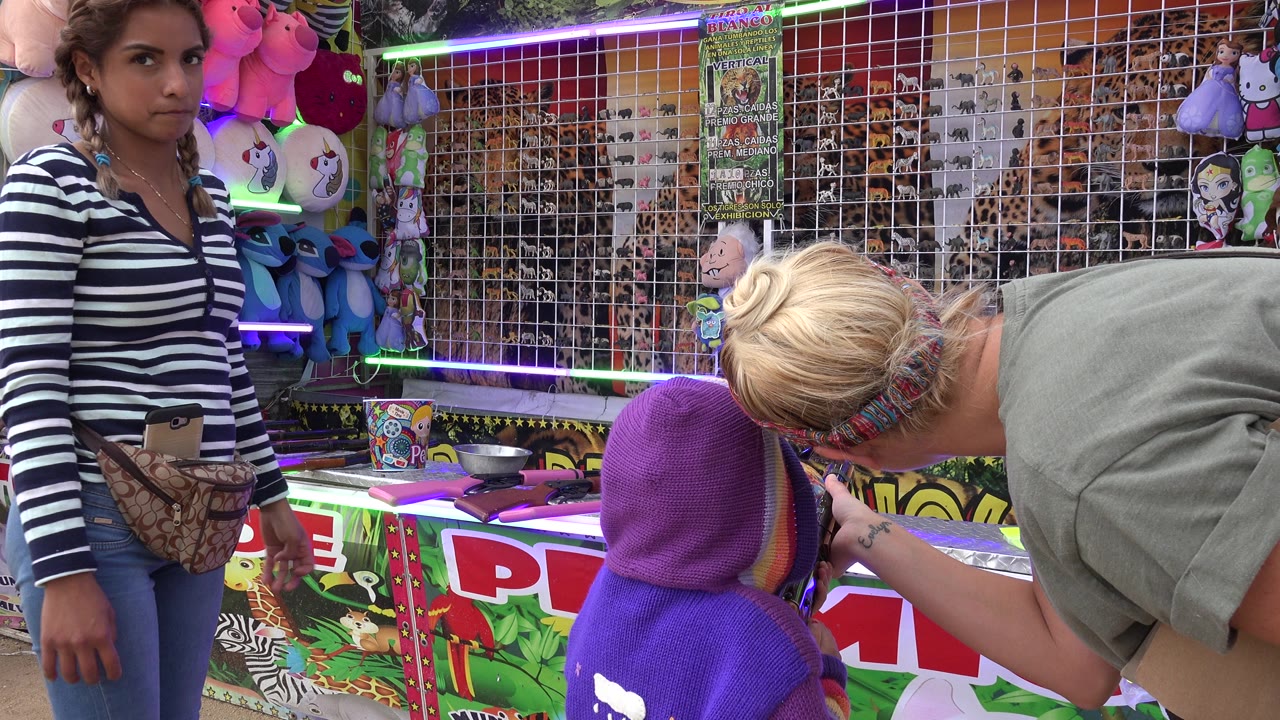 Child in a purple hood faces a carnival air rifle booth as adults stand nearby.