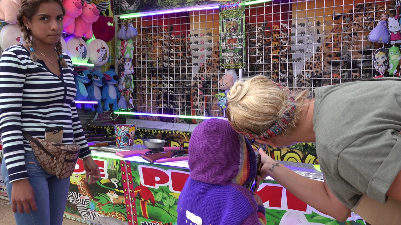 At 0:01 in San Jose del Cabo, Mexico, the child remains in front of the carnival shooting gallery as a blond adult bends near the rifle and the target grid fills the back wall.