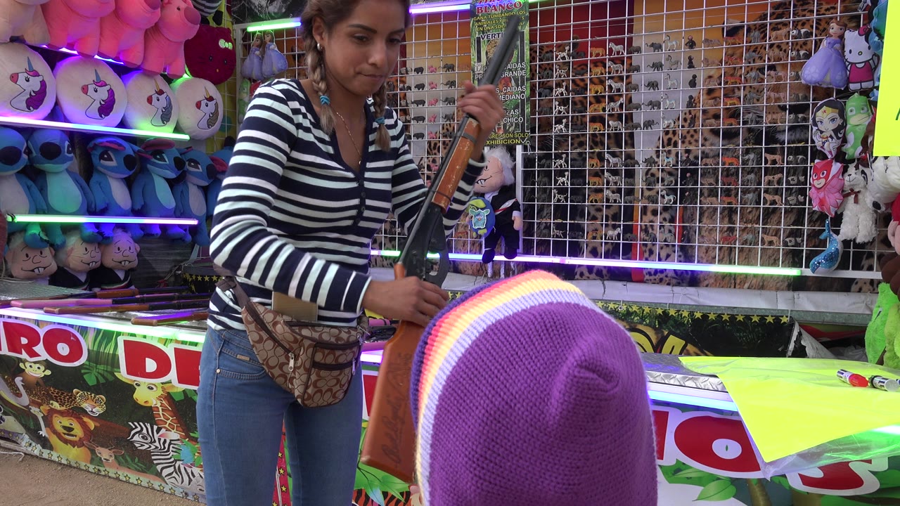 In San Jose del Cabo, Mexico, a carnival worker in a striped shirt holds a rifle prop beside a child in a purple hood at a bright prize booth.