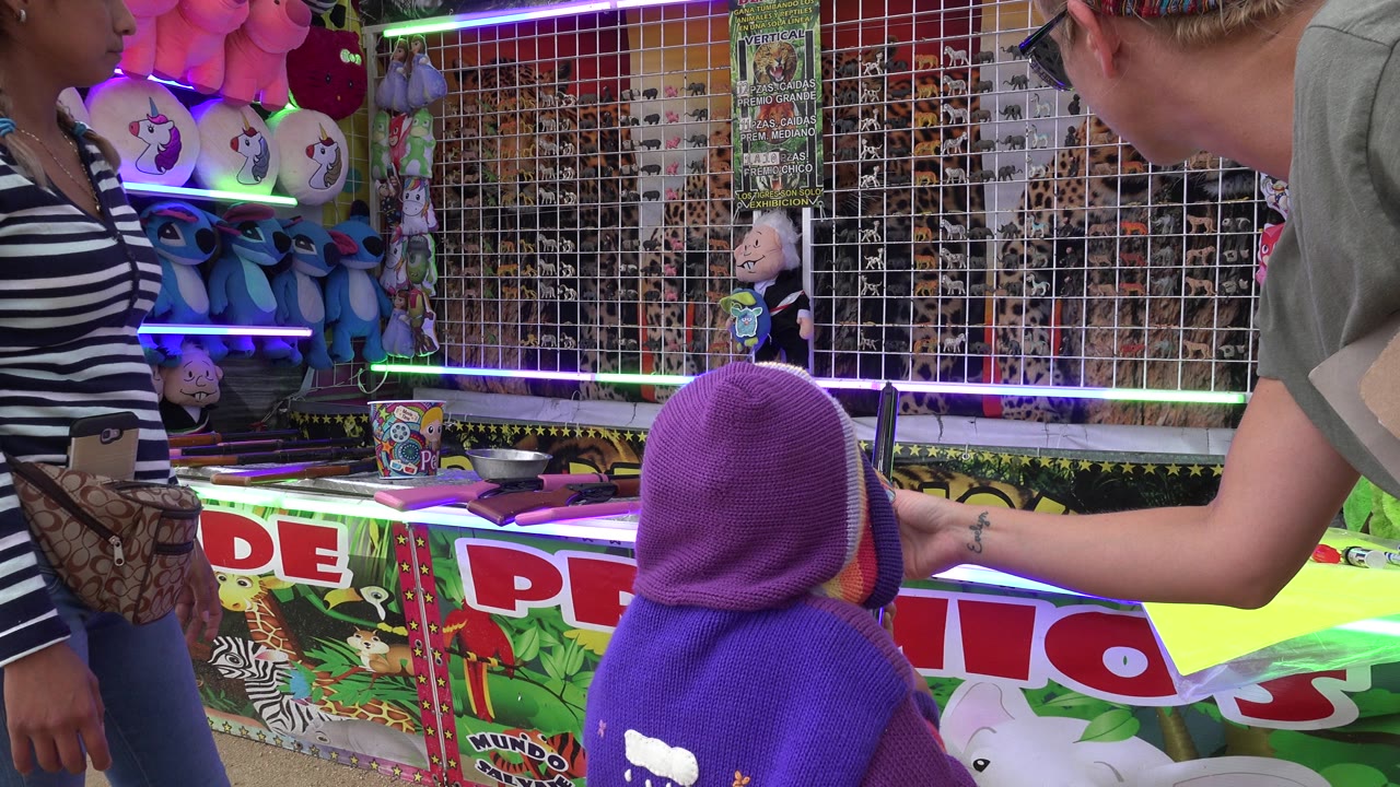 Child in a purple hood faces a carnival air rifle booth with adults nearby.