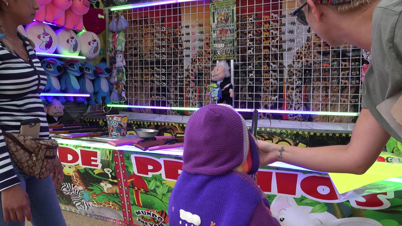 At 0:01 in San Jose del Cabo, the child remains in the foreground as an adult reaches toward the air rifle area and stuffed animal prizes line the booth behind them.