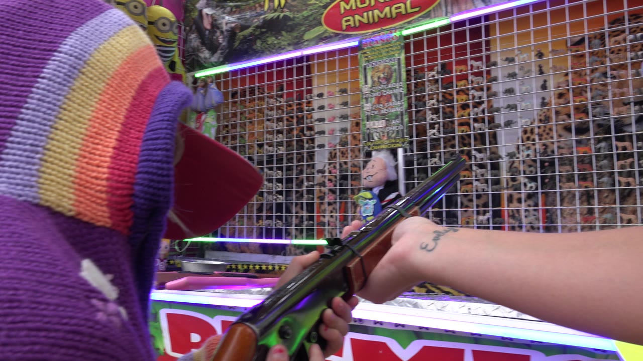 At a San Jose del Cabo fair game, a young participant in a purple striped sweater holds a carnival shooting gallery gun while facing a gridded wall of prizes.
