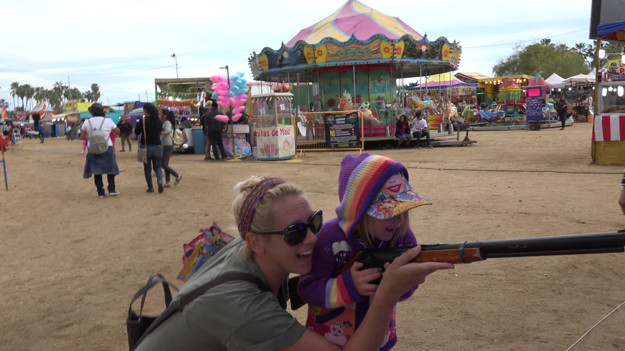 Adult woman helps a child aim a carnival rifle at an amusement park game.