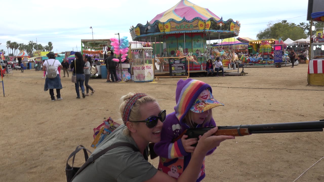 One second in, the child keeps the carnival rifle raised while the adult woman smiles close beside her, with colorful ride panels, booth signs, and people crossing the fairground in San Jose Del Cabo, Mexico.