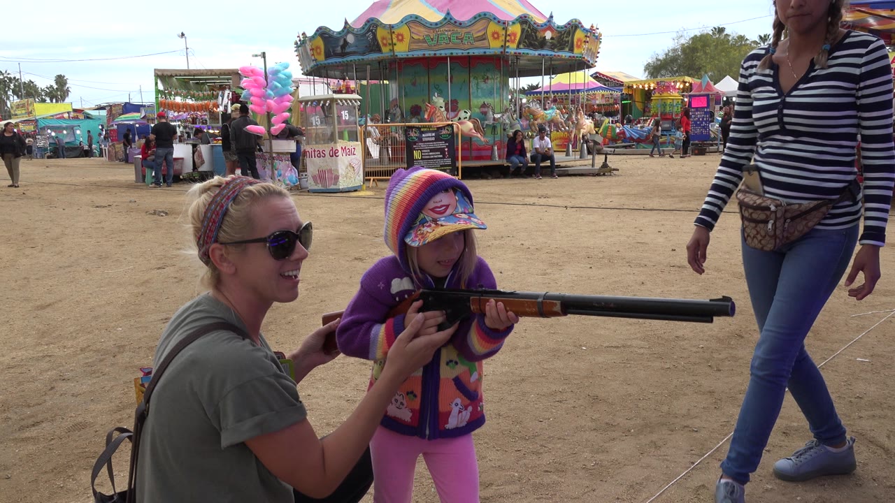 At a San Jose Del Cabo fairground, a child in a colorful hood aims a long toy rifle as a crouching woman assists and carnival booths sit behind them.