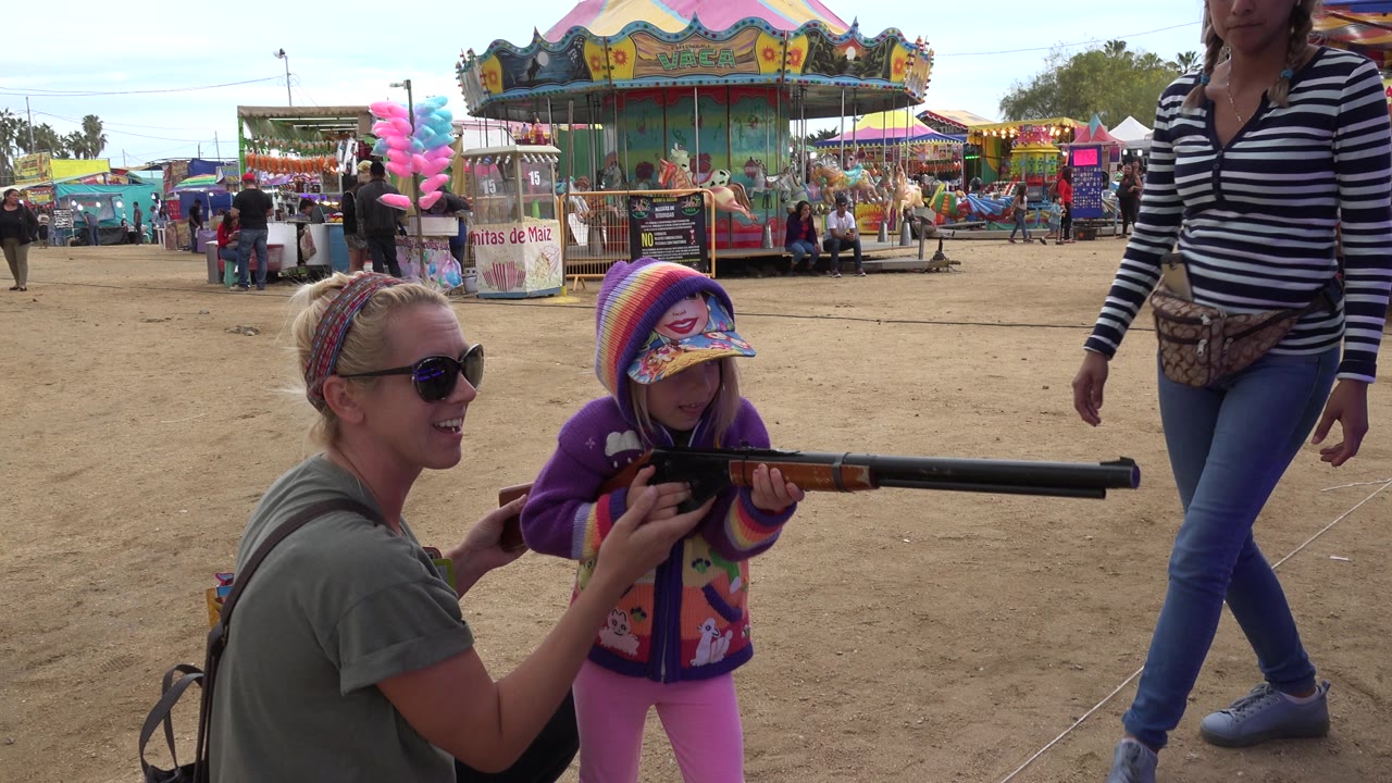 The child keeps the toy rifle raised while the crouching woman stays close, with a standing woman partly framed at right and a carousel centered in the background.