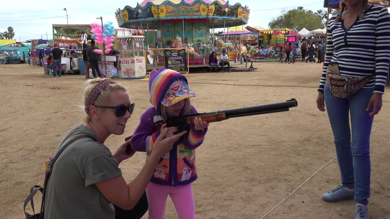 Child points a long toy rifle while two adults stand nearby at a carnival.