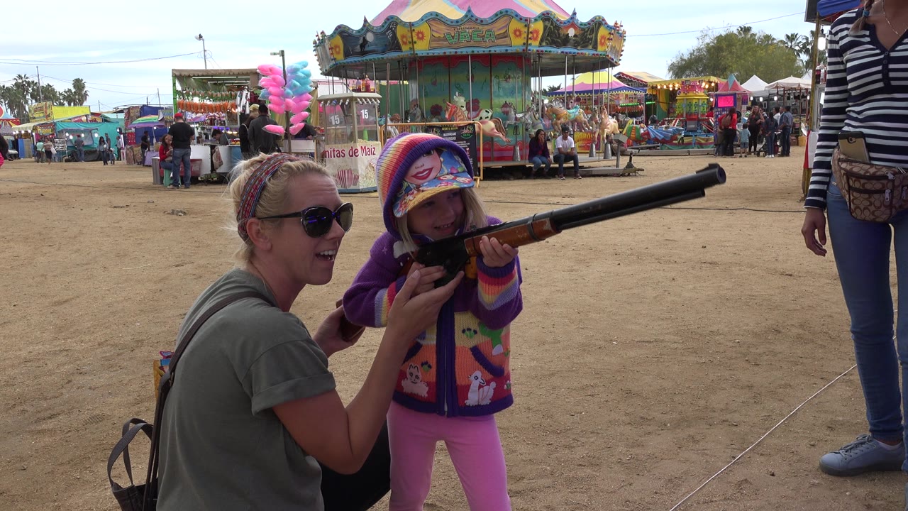 Woman helps a young girl aim a carnival rifle at a fairground game.