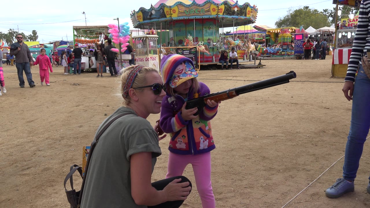Young girl aiming a rifle beside a crouching adult at a fairground.