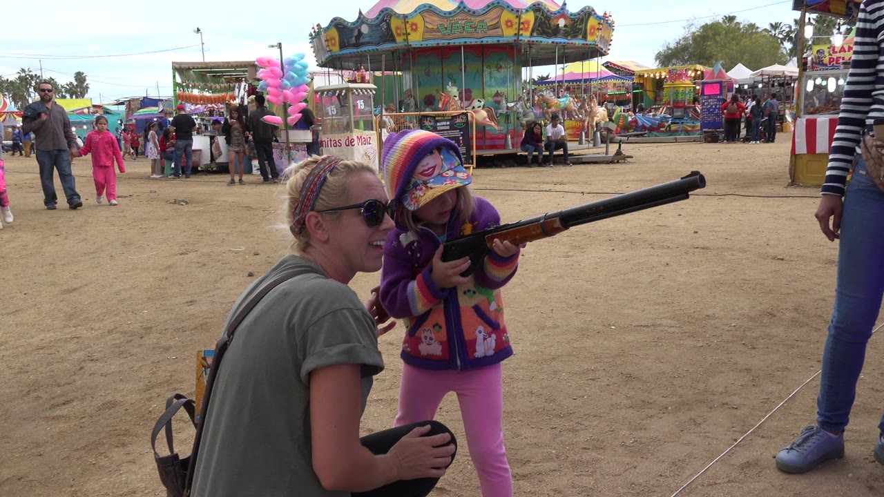 In San Jose del Cabo, Mexico, the girl holds the rifle level in front of her as the adult remains crouched close by, with carnival booths, a carousel, and passing people behind them.