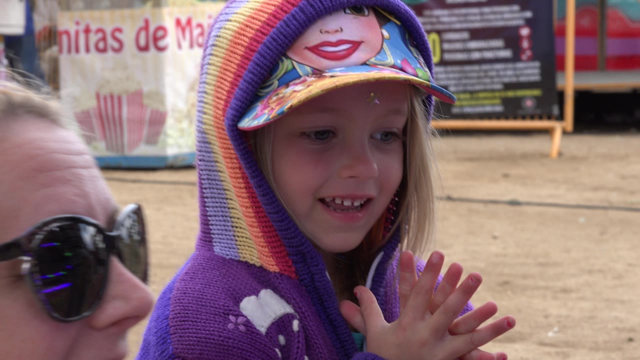 Young girl in a purple hoodie and rainbow knit hat smiles beside an adult woman in sunglasses during an outdoor San Jose del Cabo gathering.