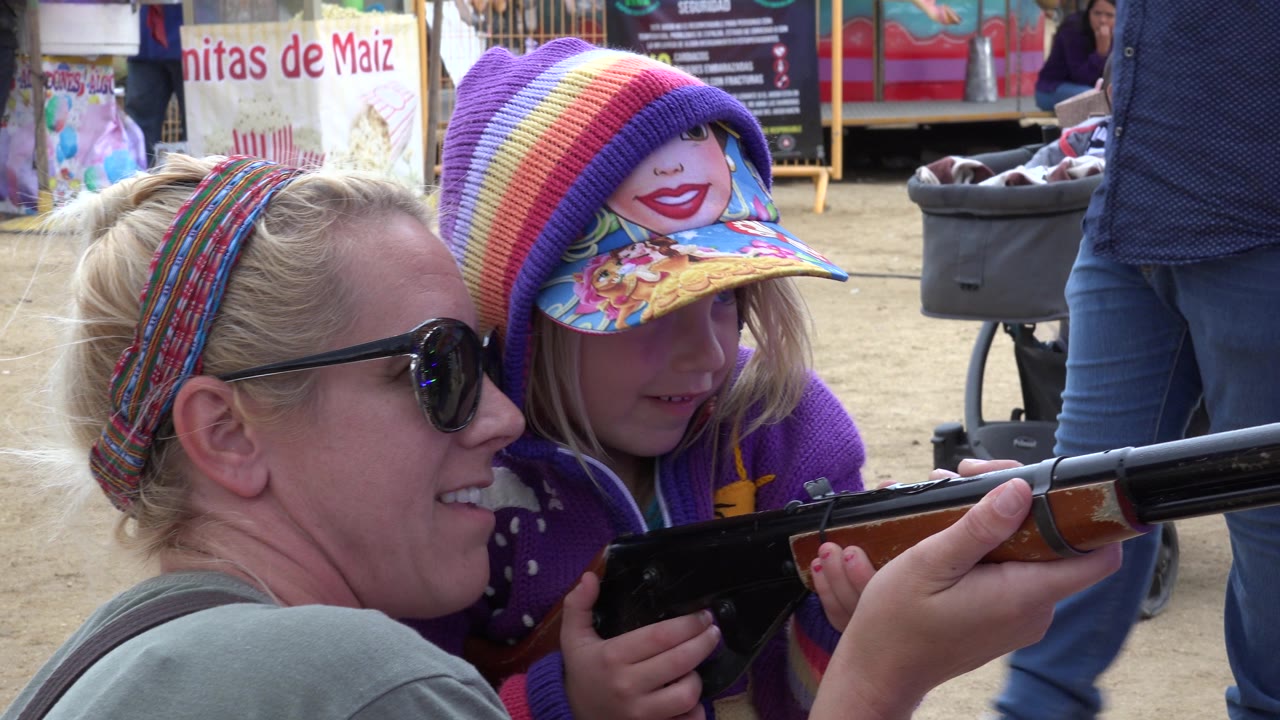 In San Jose del Cabo, Mexico, the woman smiles beside the child as both hands stay on the toy rifle, with food stand signage and people behind them.