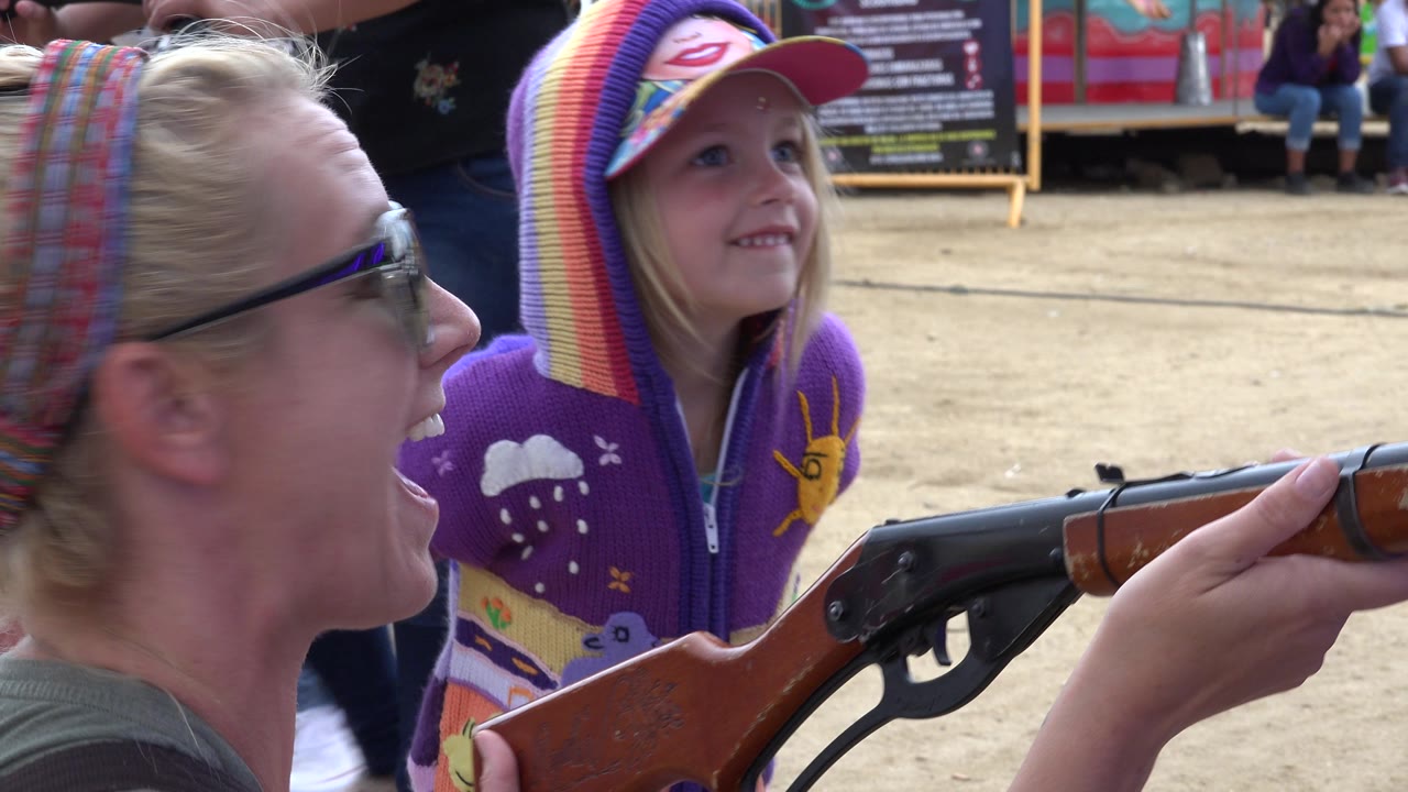 Woman in sunglasses laughs beside a girl in a rainbow hat at a carnival game.