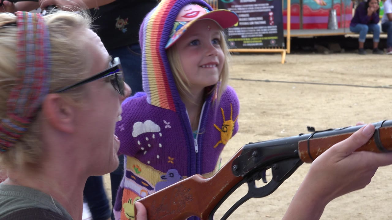 In San Jose del Cabo, Mexico, the girl in a rainbow hat smiles toward the game area while the wooden-and-black rifle-style carnival gun extends across the foreground.
