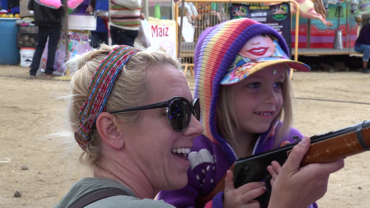 In San Jose del Cabo, Mexico, a woman in sunglasses smiles beside a young girl in a purple hood as they hold a toy rifle near food-stall signs.