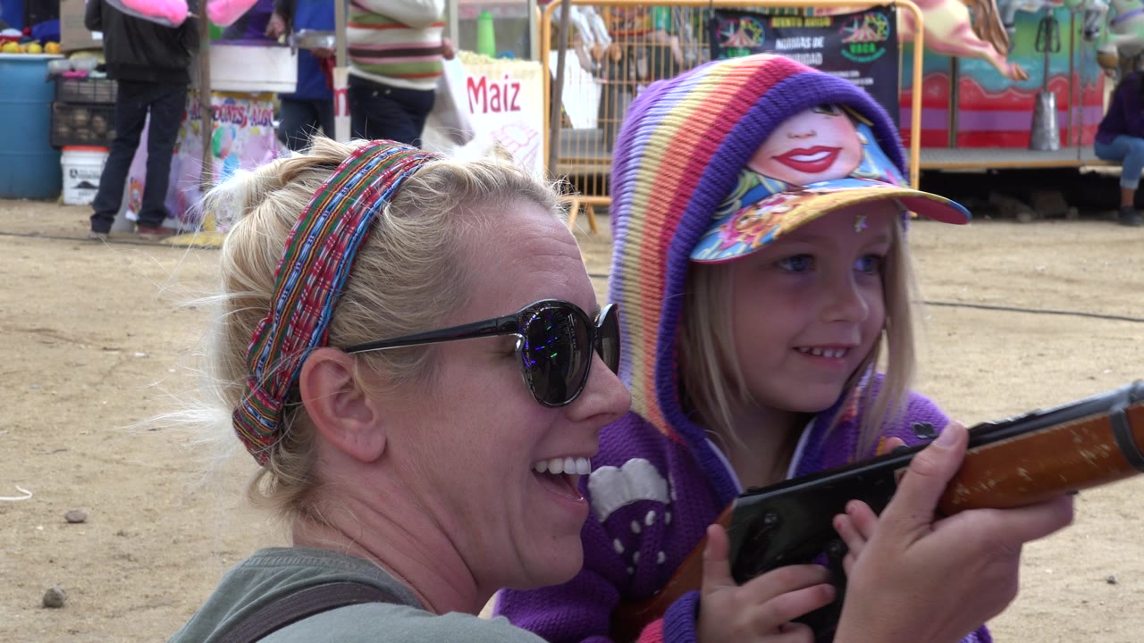 In San Jose del Cabo, Mexico, the woman grins in profile while the girl looks ahead from under a bright striped hood and cap, holding the toy rifle across the frame.