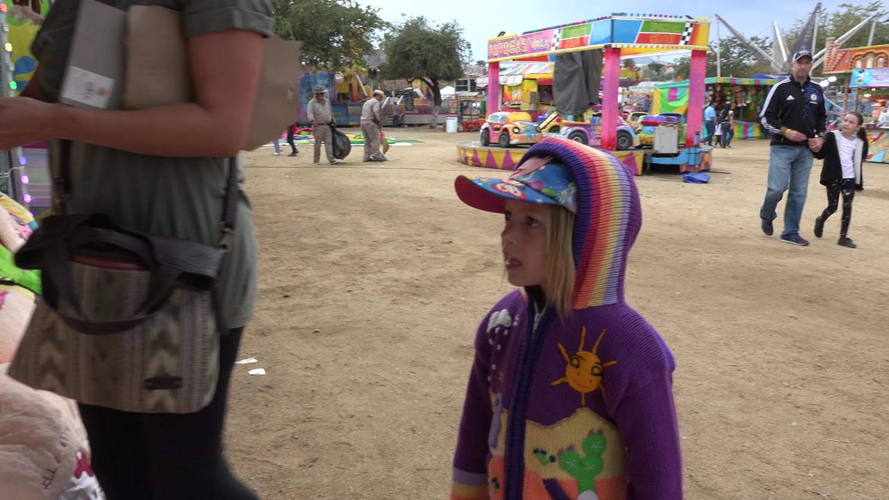 The young girl remains near the lower center of the San Jose Del Cabo fair scene as an adult woman crosses close to the camera and colorful game stalls fill the background.