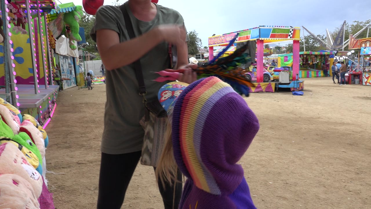 Child in striped hat faces a woman beside carnival prize toys.