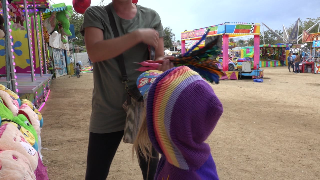 The child in the striped hat remains in the foreground while the woman stands above them near hanging plush prizes and the colorful amusement park setup in San Jose Del Cabo, Mexico.