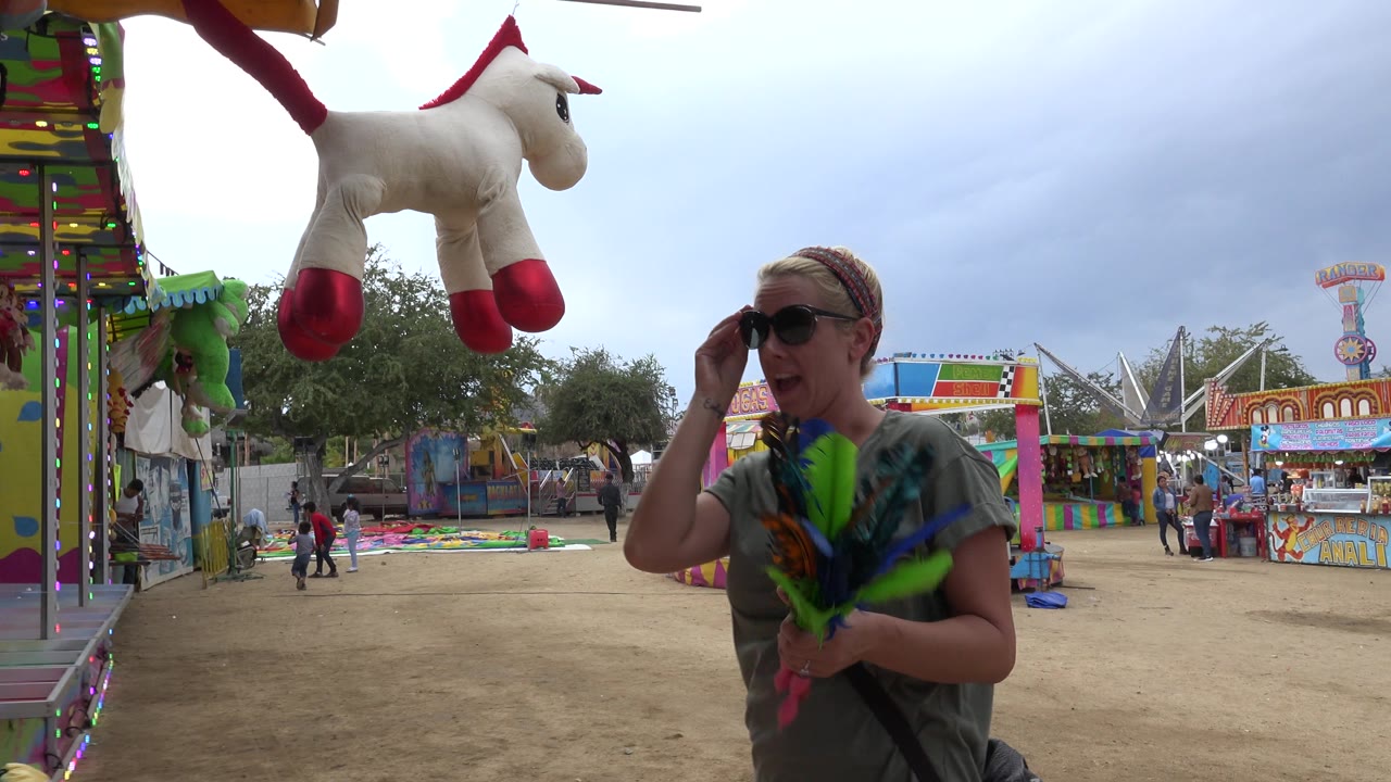 At 0:00 in San Jose del Cabo, Mexico, a woman in sunglasses holds feathered darts beside a colorful amusement booth with plush prizes overhead and fairground stalls behind her.