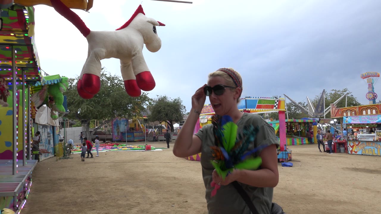 At 0:01 in San Jose del Cabo, Mexico, the woman adjusts her sunglasses while holding green and blue feathered darts near hanging stuffed animals and a bright game stall.