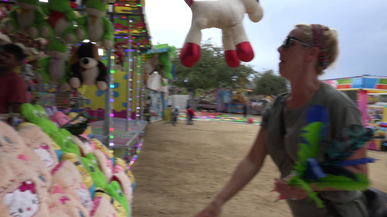 At a San Jose del Cabo carnival game, a woman holding colorful darts stands close to plush prizes while balloon targets and a fairground walkway sit behind her.