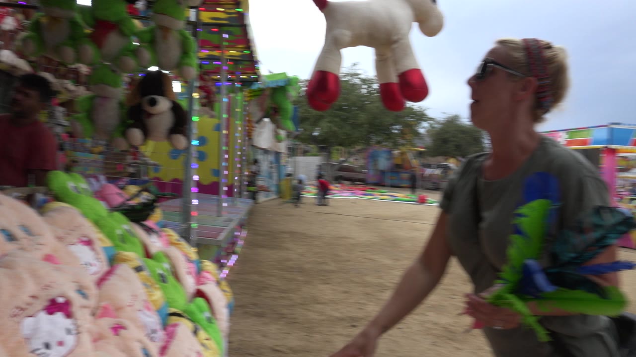 The woman remains near the booth edge as hanging stuffed animals, bright lights, and rows of prizes fill the San Jose del Cabo carnival game frame.