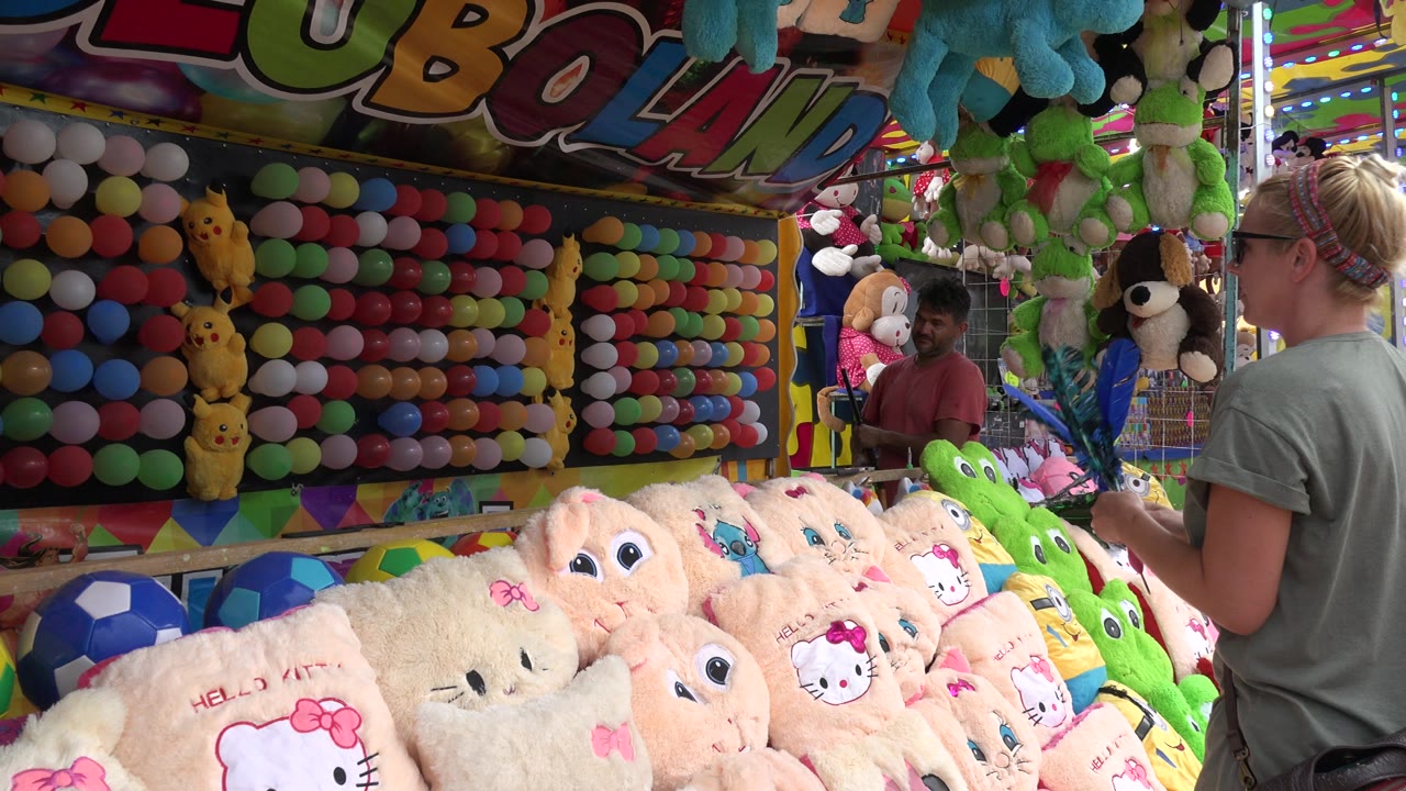 At 0:01 in San Jose del Cabo, Mexico, the woman remains at the right edge of the balloon booth while rows of balloons, pale plush toys, and a man inside the booth fill the frame.