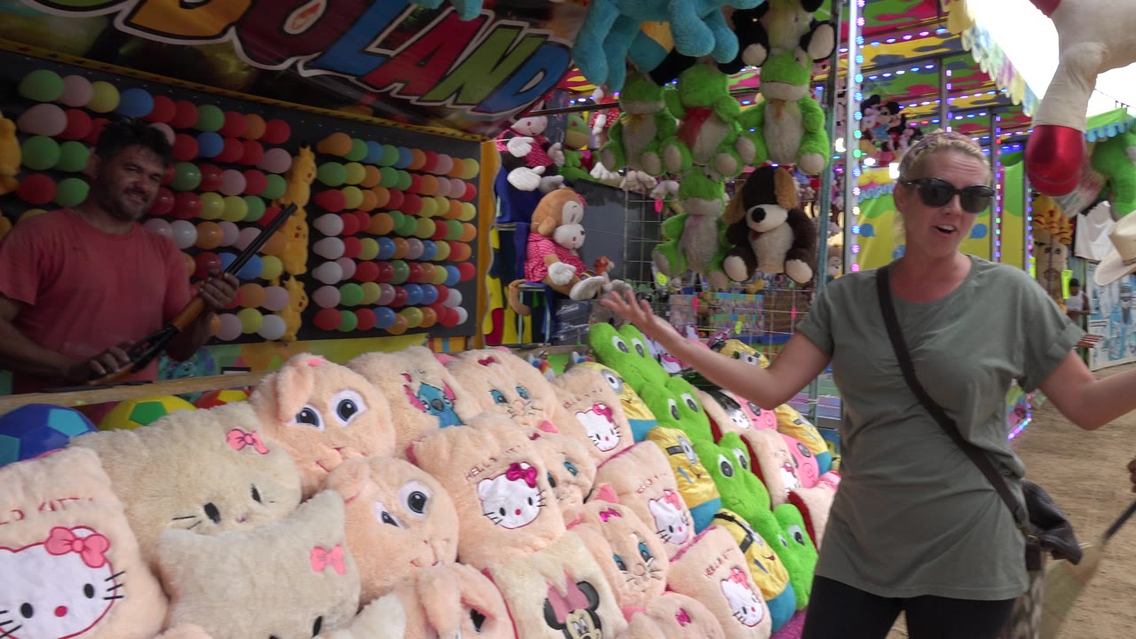 The woman gestures with both hands near a packed plush toy display in San Jose Del Cabo, Mexico, while the fair worker stands inside the booth.