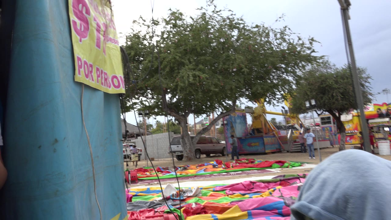 A close view in San Jose Del Cabo, Mexico shows a blue inflatable column, bright vinyl mats, trees, fencing, and a posted yellow price sign near the top edge, with no visible people in the main frame.
