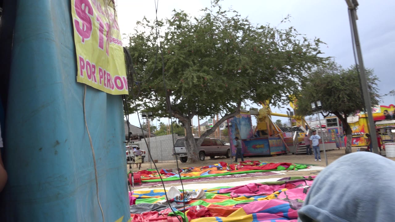 The San Jose Del Cabo frame holds on the blue inflatable column with wrinkled colorful vinyl spread across the ground, while trees, fencing, and parked vehicles remain visible in the background.